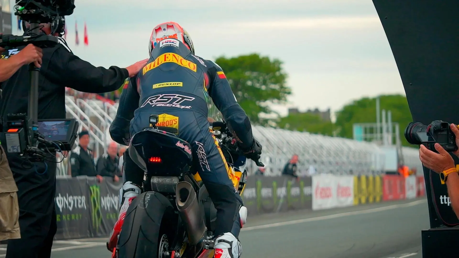 A motorcyclist prepares for a race on an open track, with a camera crew filming nearby. The sky is overcast, and the mood is tense and focused.